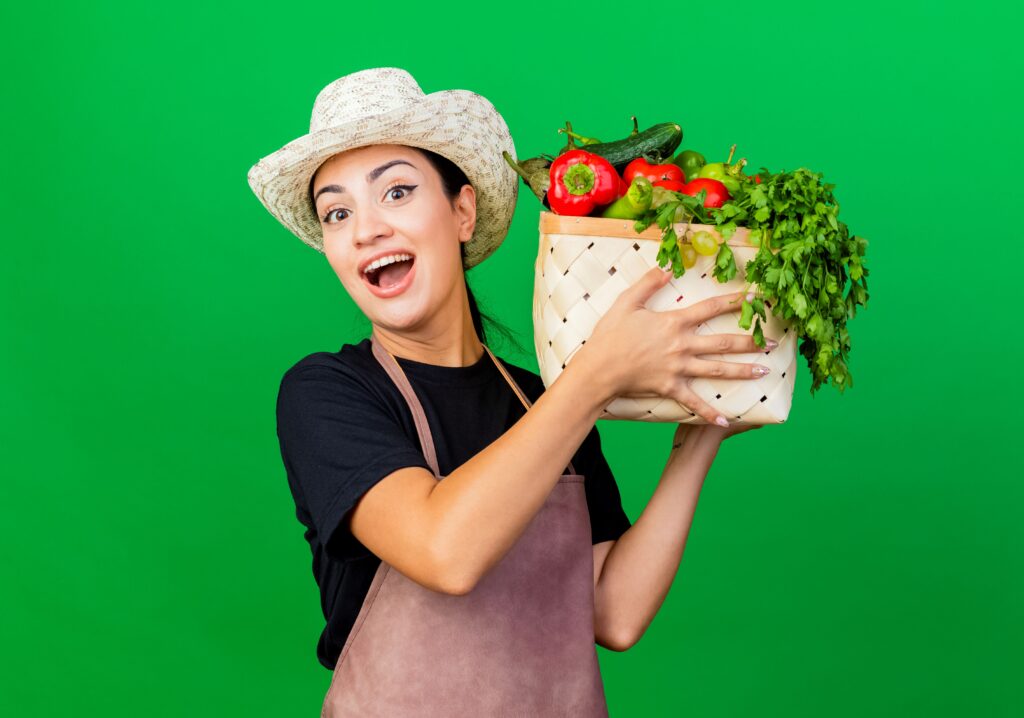 jovem-e-bela-jardineira-de-avental-e-chapeu-segurando-uma-caixa-cheia-de-legumes-olhando-para-a-frente-sorrindo-com-uma-cara-feliz-em-pe-sobre-a-parede-verde